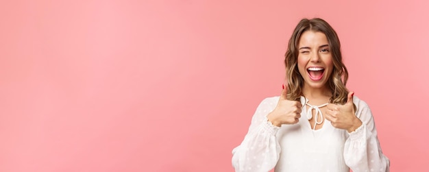 Portrait of sassy goodlooking blond girl assure all good show thumbsup and wink with classy pleased smile recommend visit place shop at online store standing in white dress pink background