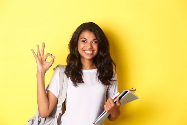 Portrait of satisfied african-american female student, smiling pleased and showing okay sign, like something good, standing over yellow background.