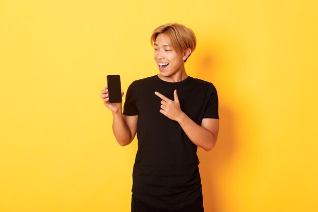 Портрет довольного азиатского парня, указывающего пальцем и смотрящего на экран смартфона с довольной улыбкой, показывающего приложение, стоящего на желтой стене
