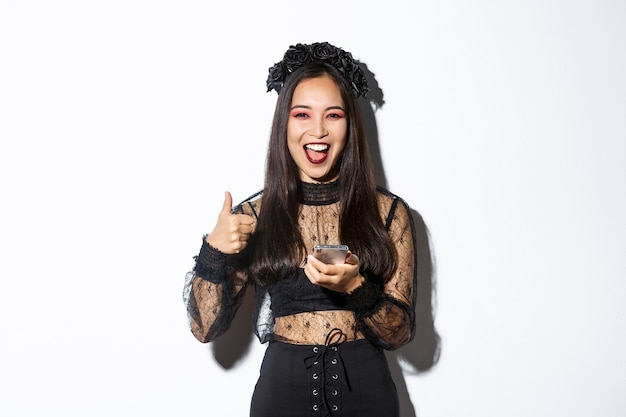 Portrait of satisfied asian woman in elegant gothic dress and black wreath showing thumbs-up while using mobile phone, standing over white background.