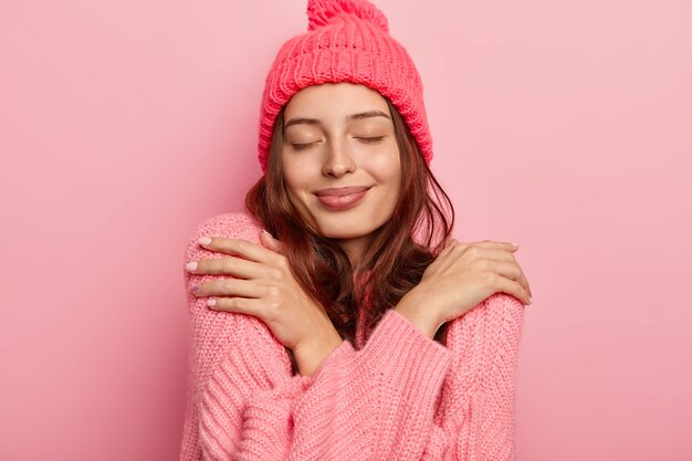 満足しているブルネットの女性の肖像画は、自分を抱きしめ、ニットの暖かいセーターで快適さを楽しんで、目を閉じて、ピンクの背景の上に隔離された新しい冬の服を購入します。