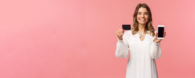 Portrait of satisfied cute blond girl in white dress showing you her credit card and mobile phone application online shopping app or bank website smiling camera standing pink background