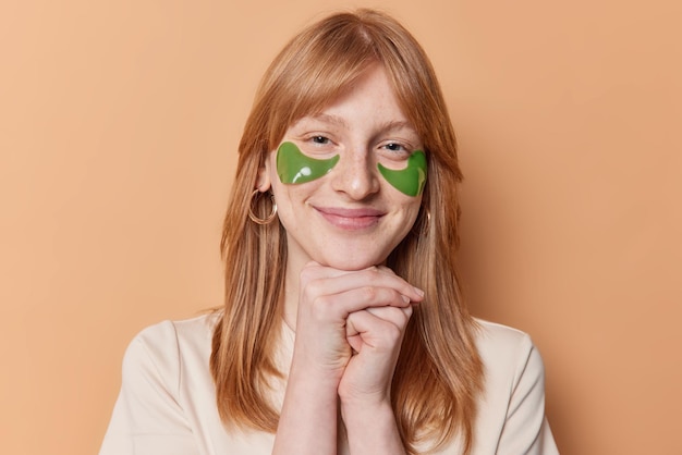 Portrait of satisfied ginger girl smiles gently keeps hands under chin applies green hydrogel patches under eyes undergoes skin care treatments dressed casually isolated over brown background