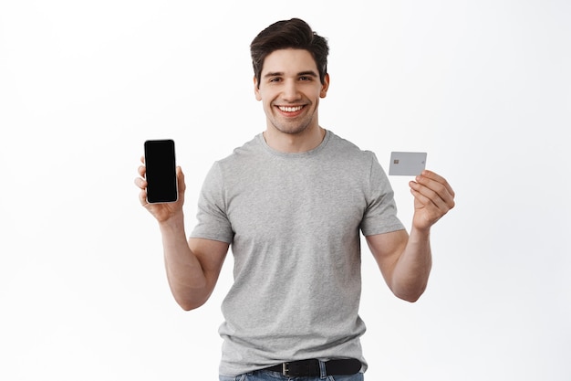 빈 스마트폰 화면과 플라스틱 신용 카드를 보여주는 만족스러운 잘 생긴 남자의 초상화, 전화 앱, 은행 및 금융 개념을 보여줍니다.