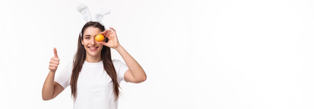 Portrait of satisfied happy young woman in rabbit ears holding one colored eggs over eye show thumbs