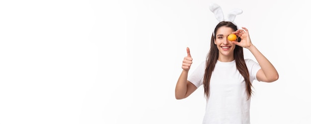 Portrait of satisfied happy young woman in rabbit ears holding one colored eggs over eye show thumbs