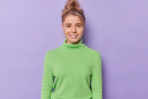 Portrait of satisfied millennial girl smiles gently has healthy skin no makeup natural beauty looks directly at camera with satisfied expression wears green poloneck isolated over purple background