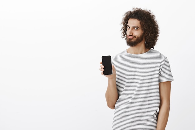 Portrait of satisfied playful Eastern bearded man with afro haircut, showing smartphone