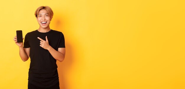 Portrait of satisfied smiling asian guy with blond hair looking amused and pointing finger at smartp