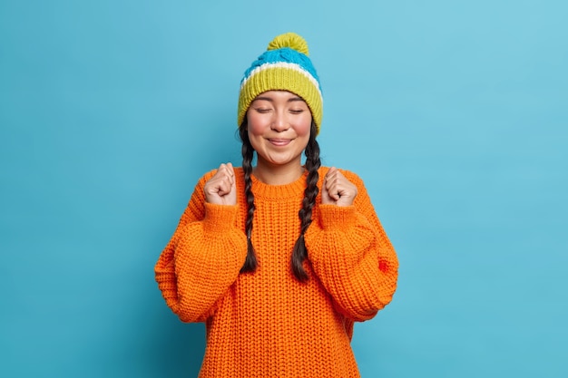 Portrait of satisfied teenage girl poses with closed eyes smiles pleasantly keeps fists clenched cannot wait until something good happen dressed in winter knitted jumper and warm hat poses indoor