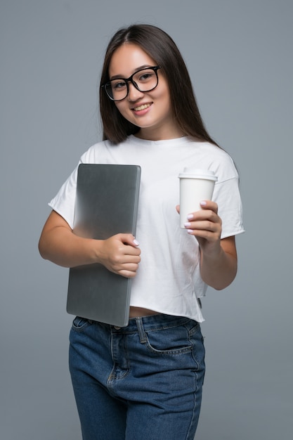 걷고 회색 배경 위에 카메라를 보면서 커피와 노트북 컴퓨터의 컵을 들고 만족 된 젊은 아시아 여자의 초상화