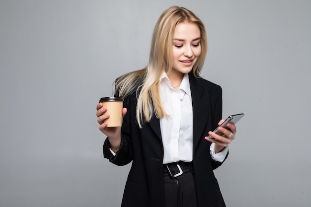 Portrait of a satisfied young business woman using mobile phone while holding cup of coffee to go isolated