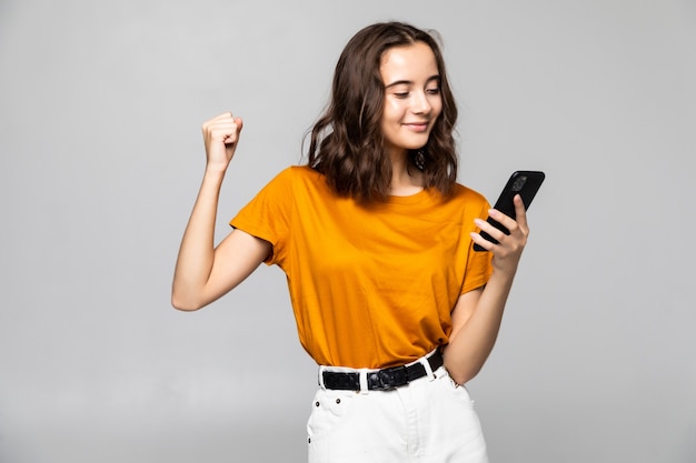 Portrait of a satisfied young woman holding mobile phone and celebrating isolated over gray 