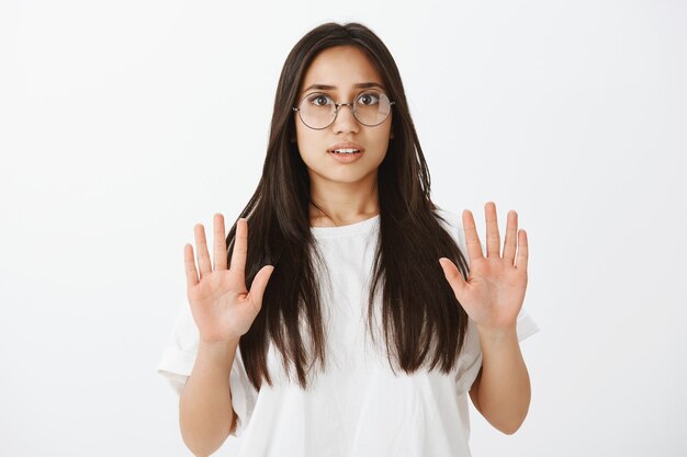 Portrait of scared beautiful young female student in glasses and white t-shirt, raising palms high in surrender and making frightened intense expression, asking guy chill or relax while arguing