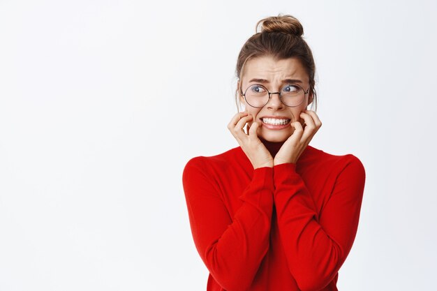 Portrait of scared businesswoman biting fingernails and looking left frightened, panicking and staring with fear at copy space, standing against white wall