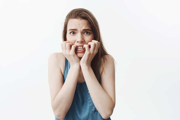 Portrait of scared good-looking young woman with dark long hair in blue clothes gnawing hands,  with frightened expression, being nervous and unconfident about next exam.