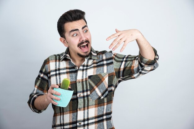 Portrait of scared man keeping little cactus in blue pot over white wall.