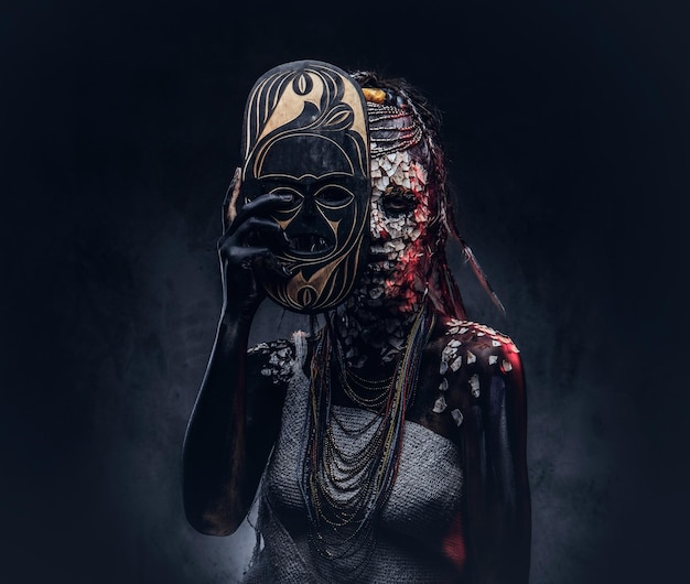 Portrait of a scary African shaman female with a petrified cracked skin and dreadlocks, holds a traditional mask on a dark background. Make-up concept.