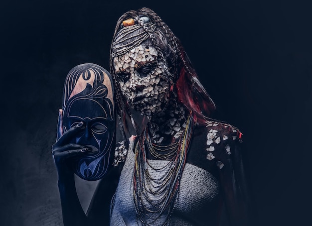石化したひびの入った肌とドレッドヘアを持つ恐ろしいアフリカのシャーマンの女性の肖像画は、暗い背景に伝統的なマスクを保持しています。メイクアップのコンセプト。