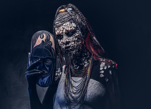 Portrait of a scary African shaman female with a petrified cracked skin and dreadlocks, holds a traditional mask on a dark background. Make-up concept.