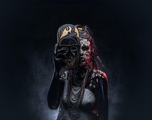 Ritratto di una donna sciamana africana spaventosa con una pelle screpolata pietrificata e dreadlocks, tiene una maschera tradizionale su uno sfondo scuro. concetto di trucco.