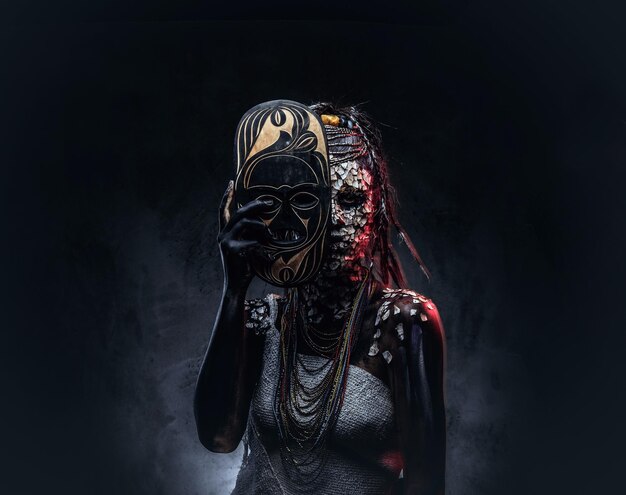 Portrait of a scary African shaman female with a petrified cracked skin and dreadlocks, holds a traditional mask on a dark background. Make-up concept.