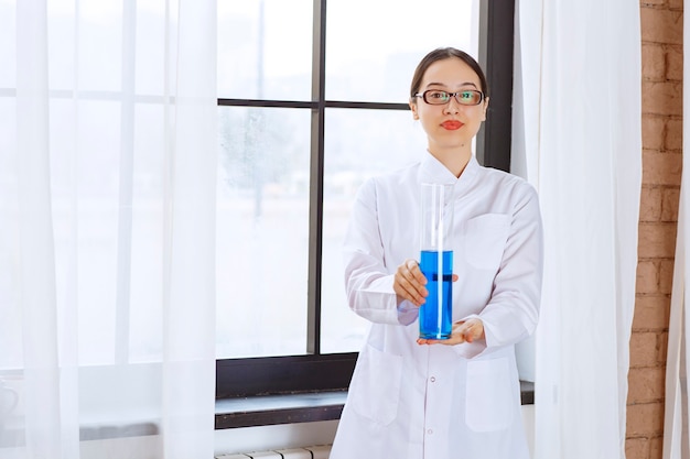 Portrait of scientist woman in lab coat holding chemical blue liquid .