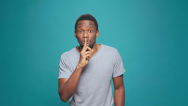 Portrait of secretive man doing hush sign in front of camera, putting index finger over mouth and lips. Male model showing mute and silence gesture, asking for privacy about confidential gossip.