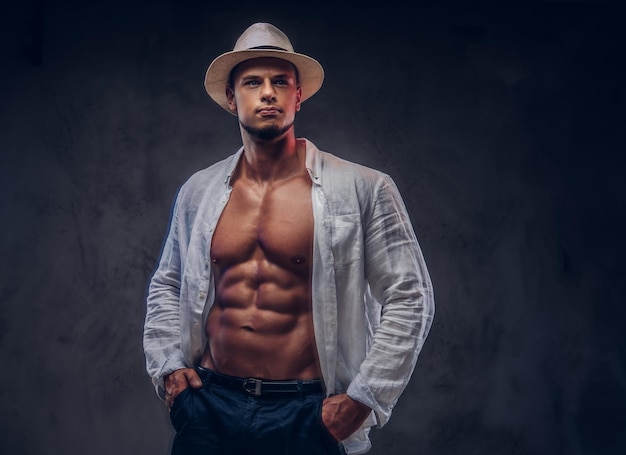 Portrait of a seductive sexy handsome guy with muscular body in an unbuttoned white shirt and panama hat. Isolated on dark background.