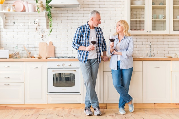 Portrait of senior couple holding wineglasses in hand looking at each other standing in kitchen