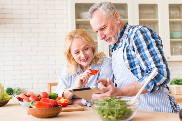 台所でサラダを準備しながらデジタルタブレットを見て年配のカップルの肖像画
