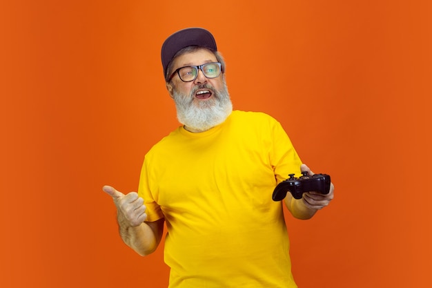 Portrait of senior hipster man using devices, gadgets isolated on orange studio background. Tech and joyful elderly lifestyle concept.