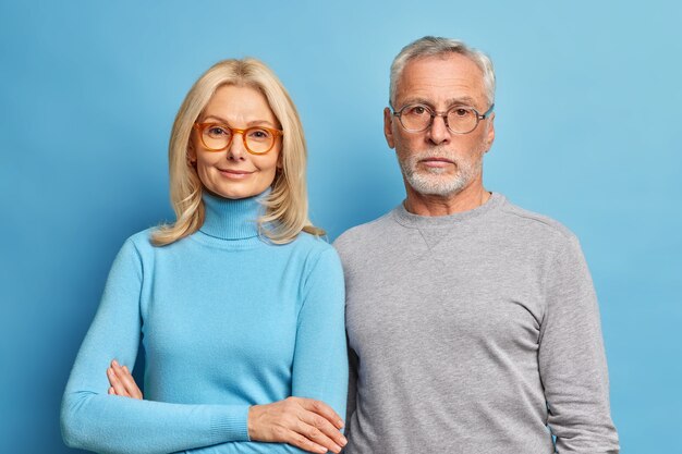Portrait of senior husband and wife pensioners stand closely to each other dressed in casual clothes and spectacles enjoy sweet moments of being together or retirement isolated over blue wall