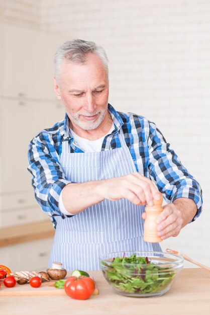 Портрет старшего человека, добавив перец с мельницей в зеленый салатник на деревянный стол