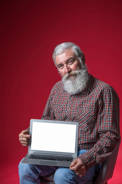 Portrait of a senior man sitting on chair showing laptop with blank white screen against red background