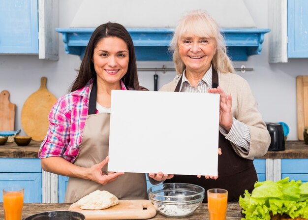 Portrait of senior mother and her young daughter holding blank white card standing in the kitchen