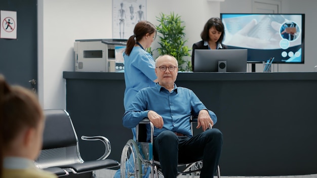 Portrait of senior wheelchair user sitting in waiting room area at hospital reception. Retired man with impairment and health condition, dealing with chronic disability in facility lobby.