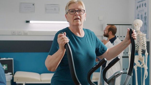 Portrait of senior woman doing physical therapy on stationary bicycle, using electrical bike to cure mechanical disorders. Elder patient doing rehabiltation exercise to increase mobility.