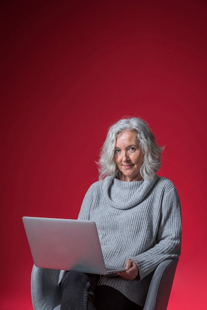 Portrait of a senior woman sitting on armchair with laptop looking to camera against red background