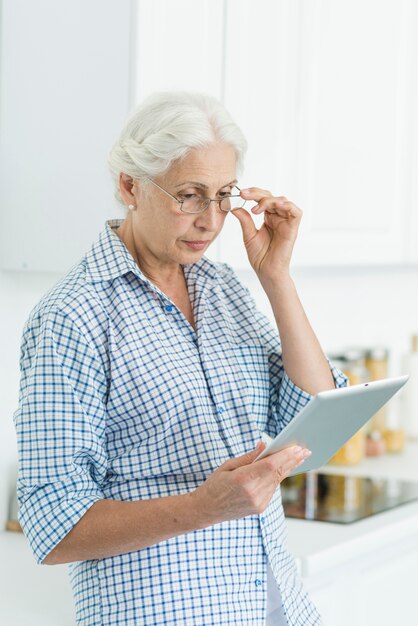 Portrait of senior woman standing in kitchen looking at digital tablet