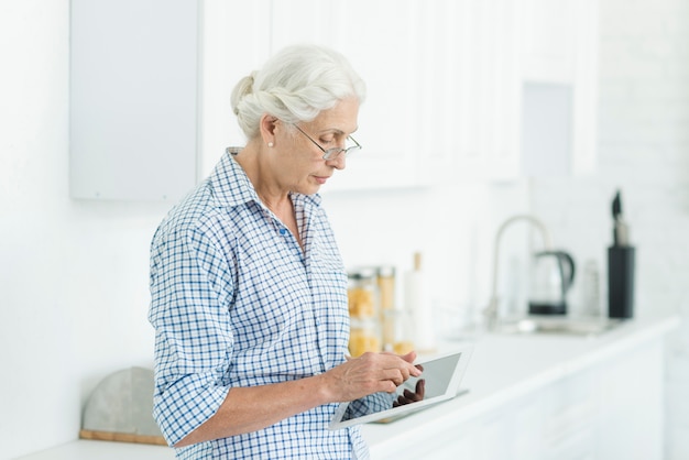 Портрет пожилой женщины, стоя в кухне с помощью цифровой планшеты