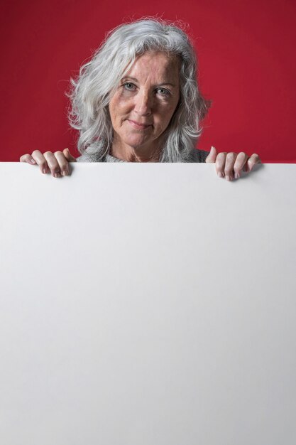 Portrait of a senior woman with grey hair standing behind the white placard against red background