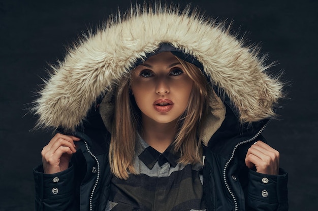 portrait of a sensual blonde girl in a winter jacket and fur hood, standing in a studio. isolated on a dark background.