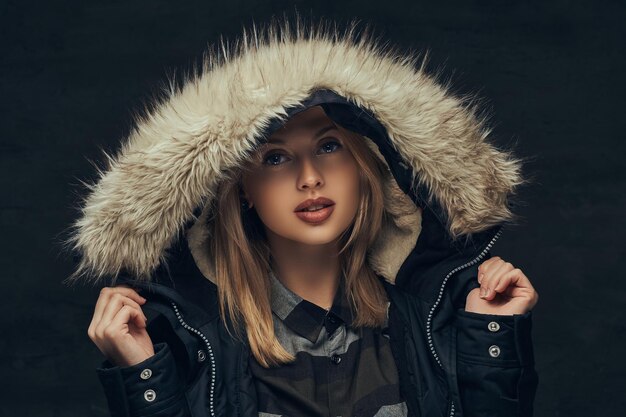 Portrait of a sensual blonde girl in a winter jacket and fur hood, standing in a studio. Isolated on a dark background.