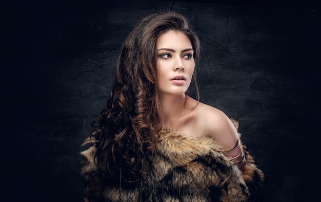 Portrait of a sensual brunette female dressed in a warm fur coat on grey background in a deep shadow.