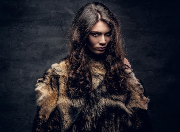 Portrait of a sensual brunette female dressed in a warm fur coat on grey background in a deep shadow.