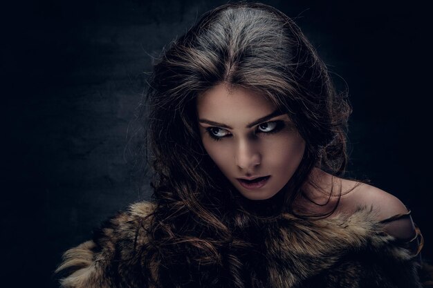 Portrait of a sensual brunette female dressed in a warm fur coat on grey background in a deep shadow.
