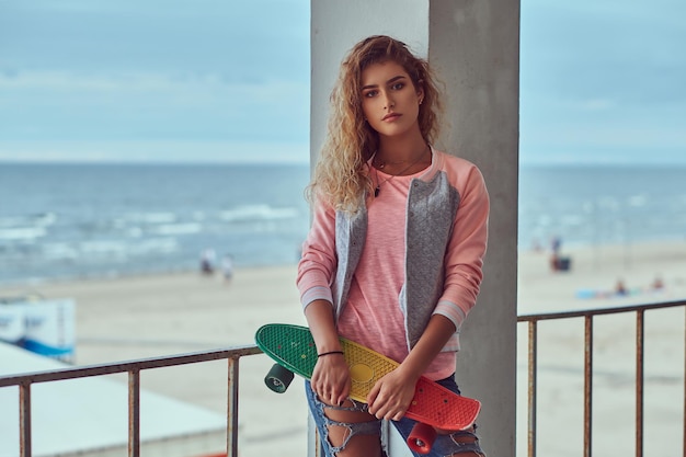 Ritratto di una ragazza sensuale con i capelli biondi vestita con una giacca rosa in piedi vicino a un guardrail contro una costa del mare.