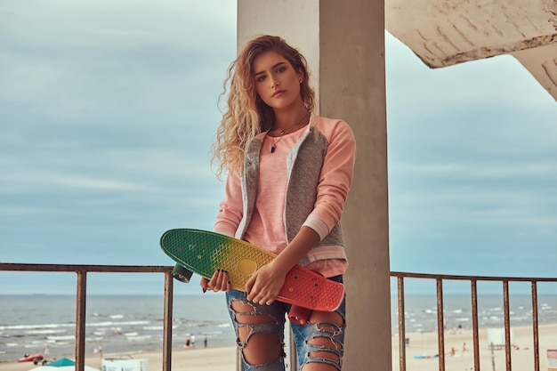 Portrait of a sensual young girl with blonde hair dressed in a pink jacket standing near a guardrail against sea coast.