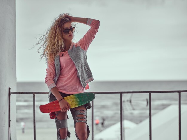 Portrait of a sensual young girl with blonde hair in sunglasses dressed in a pink jacket standing near a guardrail against black and white sea coast background.